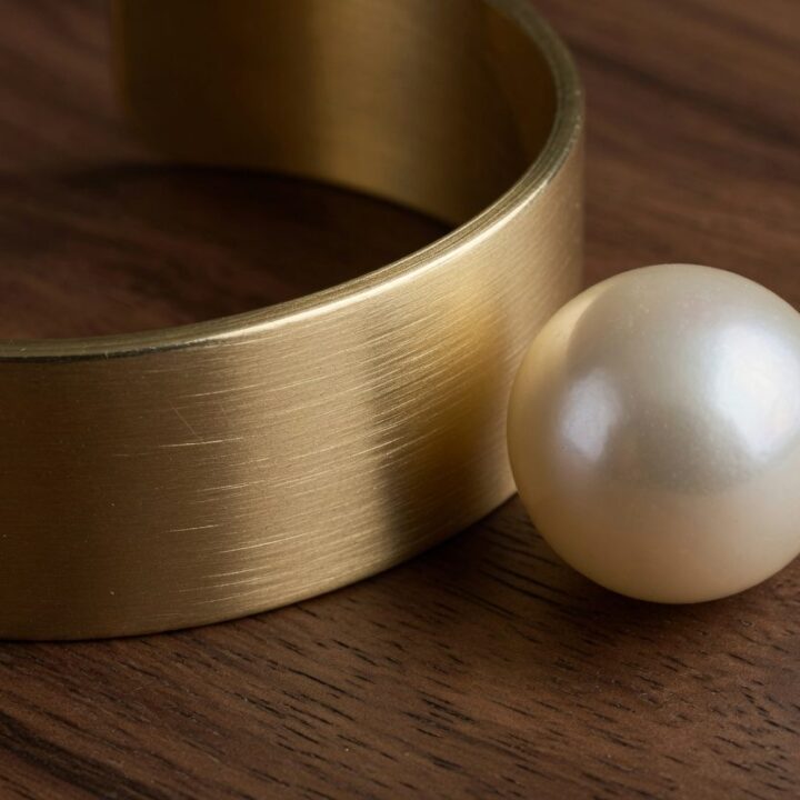 A brushed gold cuff with a loose pearl beside it, photographed on dark wood.